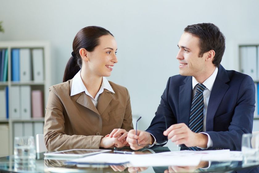 Two professionals smiling and talking across a glass table in a bright office