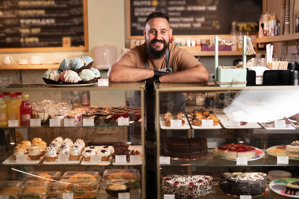 Bakery display case with assorted pastries and a smiling man leaning behind the counter