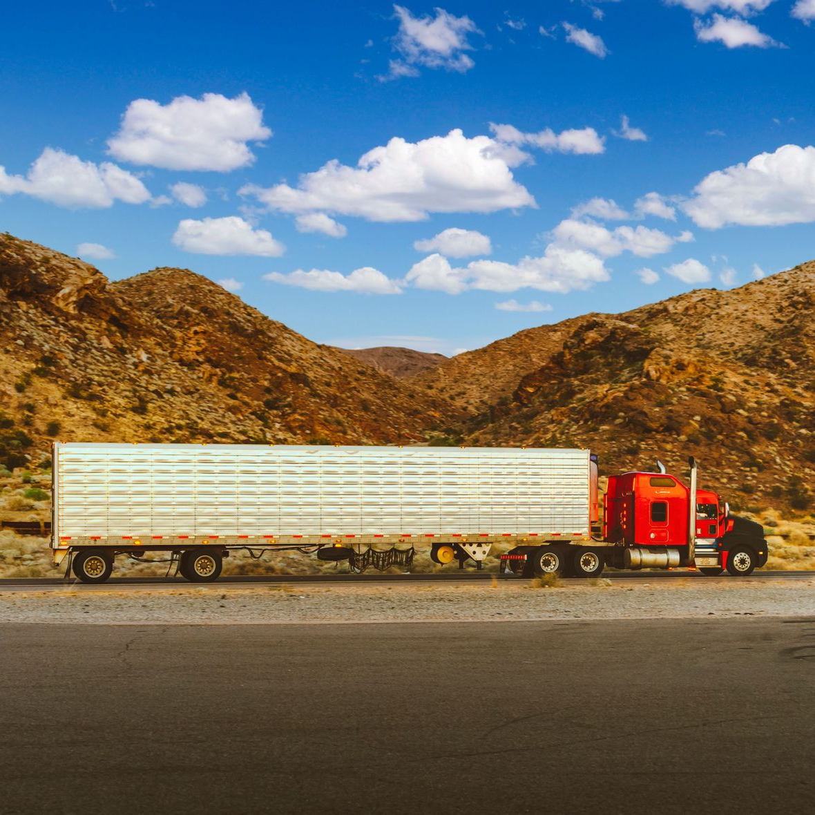 Red semi-truck with a long white trailer parked beside rocky hills under a blue sky with clouds
