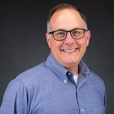 Smiling man in glasses wearing a blue patterned button-down shirt on a dark gray background