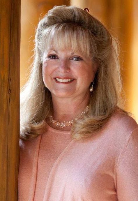 Smiling woman in a pink sweater, standing indoors near a window with warm sunlight.