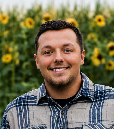 Smiling man in a blue plaid shirt standing in front of a sunflower field