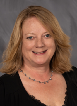 Portrait of a smiling woman with blonde hair wearing a black top and necklace against a gray backdrop