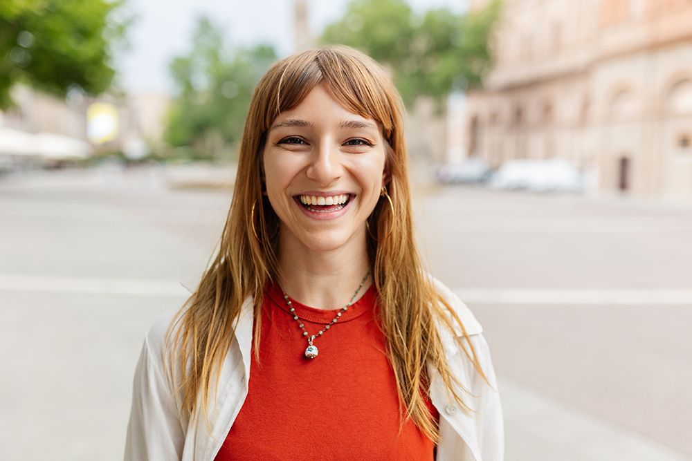 Woman with long red hair and bangs, smiling widely outside on a sunny day, wearing a red shirt and white jacket.