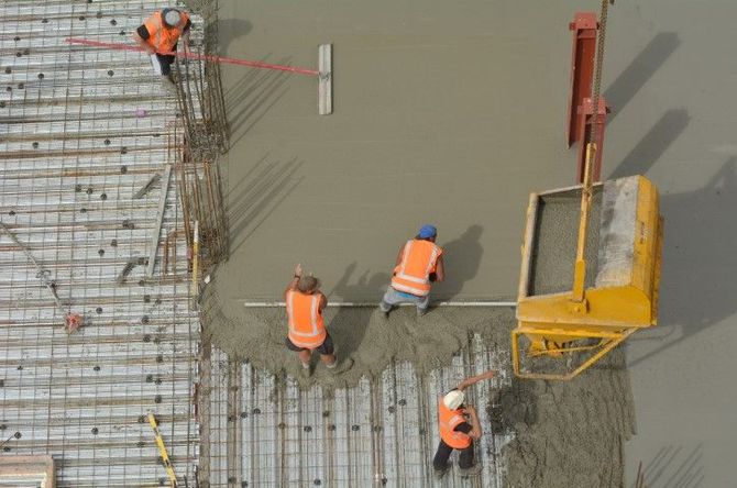 Construction workers in orange vests pour concrete beside rebar, with a yellow boom on site
