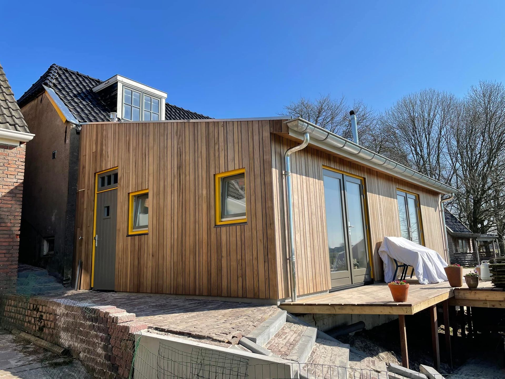 Een moderne, met hout beklede aanbouw met grote ramen en een schuin dak, naast een ouder bakstenen huis.