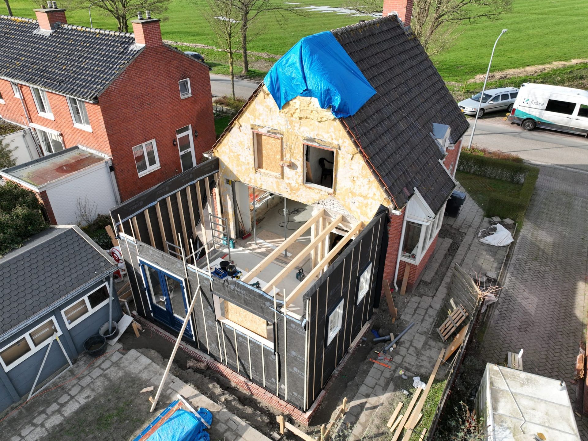 Huis in renovatie met zichtbare dakconstructie en blauw zeil erop, gezien van bovenaf.