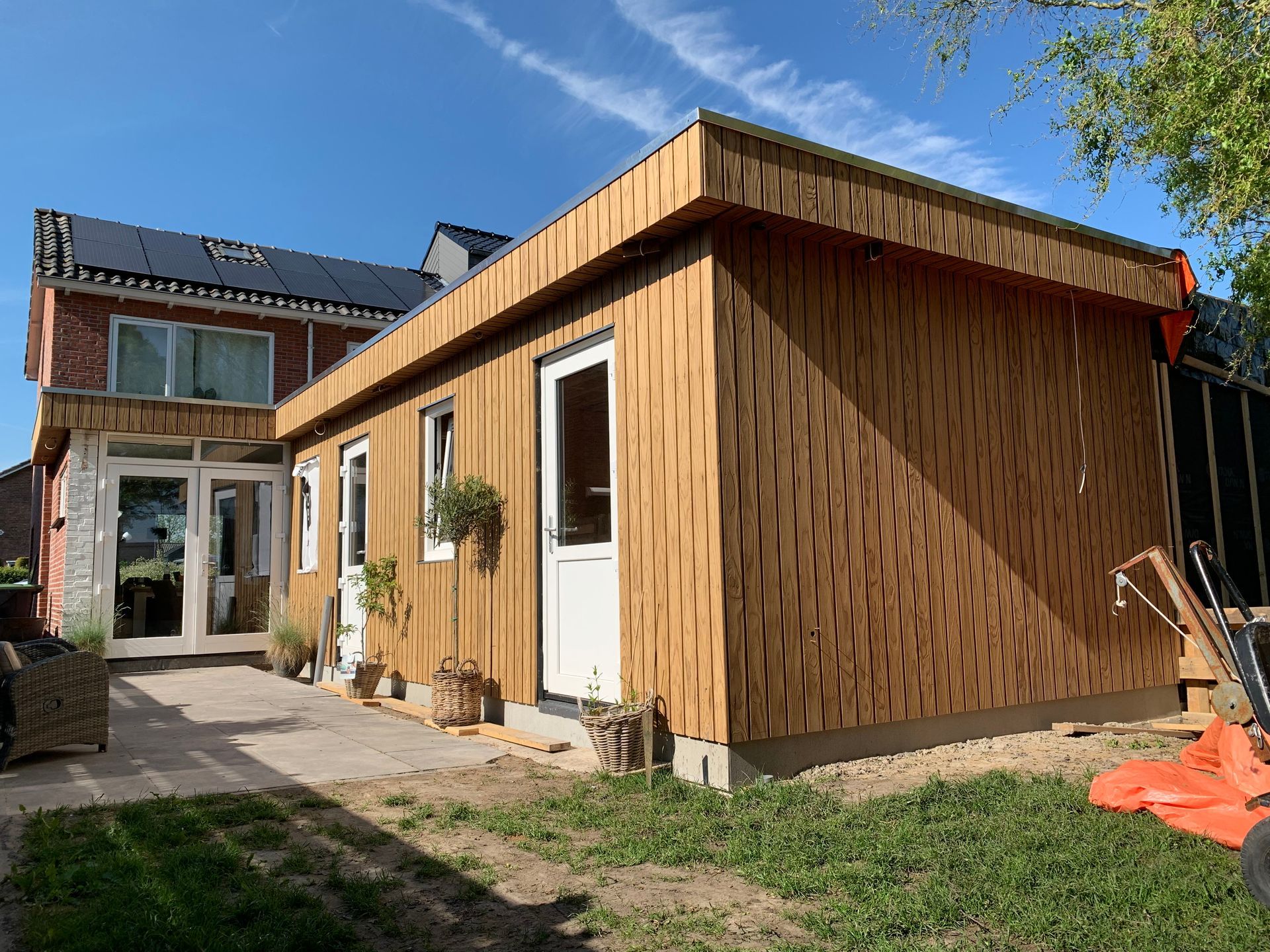 Moderne, met hout beklede aanbouw aan een huis met een terras, een gazon en een blauwe hemel.