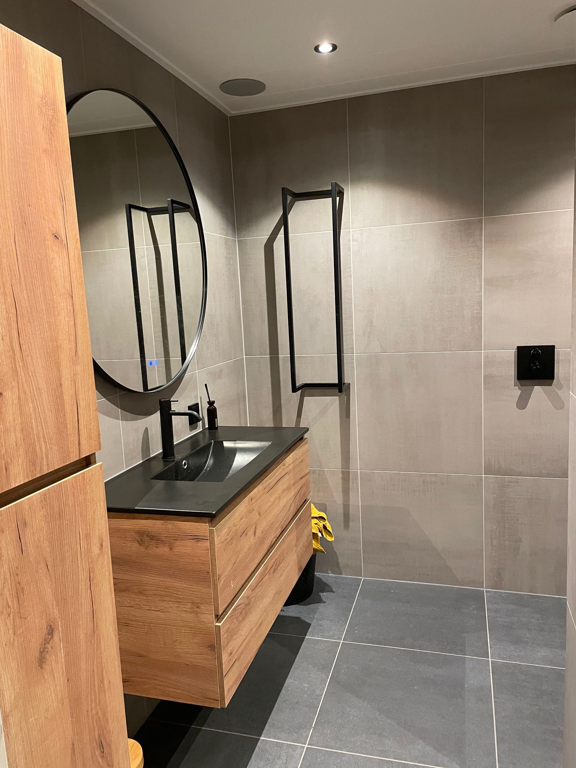 Moderne badkamer met houten badmeubel, ronde spiegel, zwarte wastafel en grijze tegelwanden.