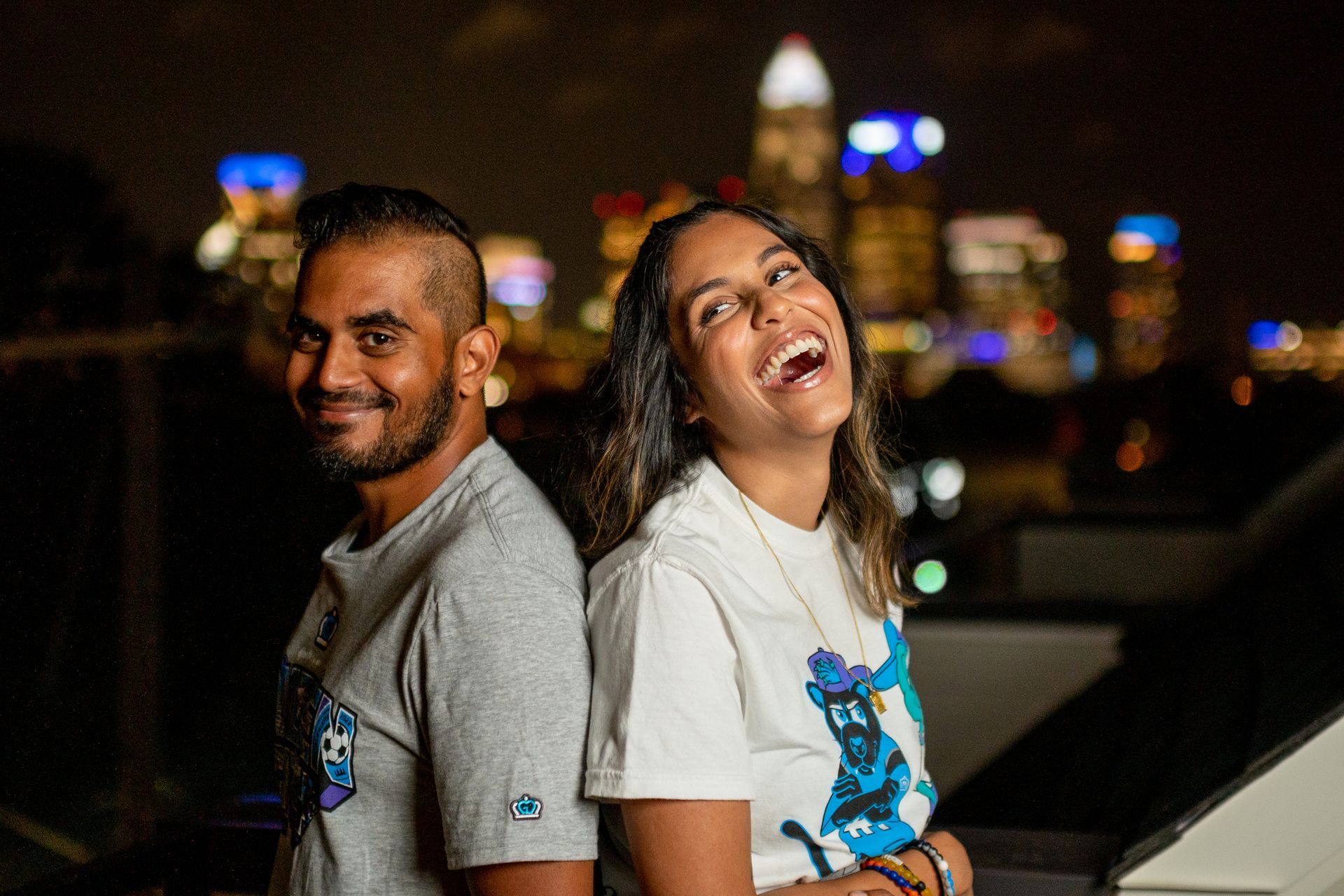 A man and a woman are standing back to back in front of a city skyline at night.