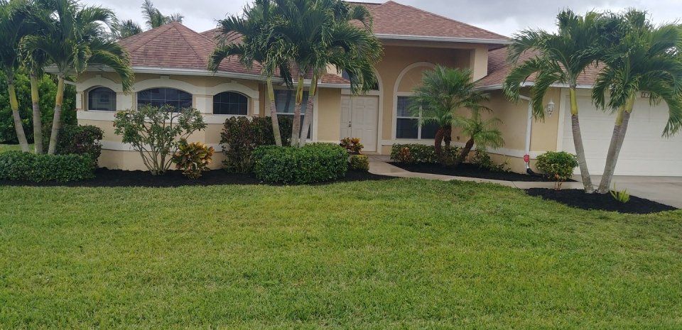 A beige house with a brown roof and palm trees in front. Green grass and black mulch are visible.
