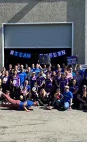 A large group wearing blue shirts poses in front of a warehouse with a 