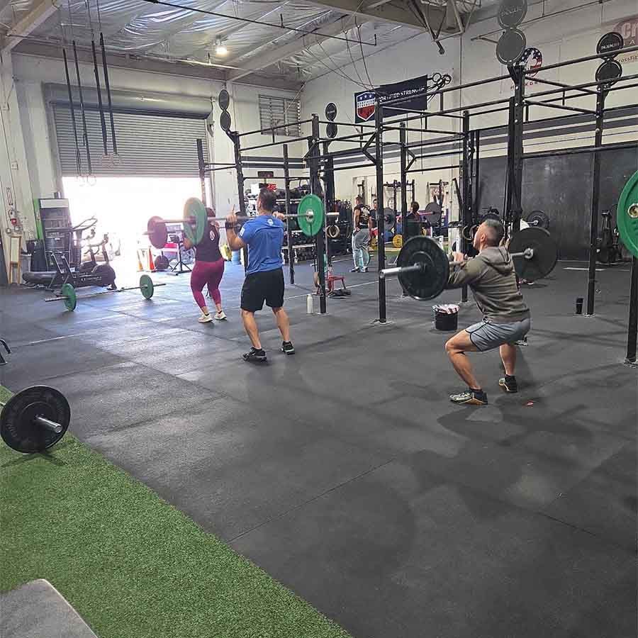 Three people perform barbell squats in a gym setting with workout rigs and equipment in the background.