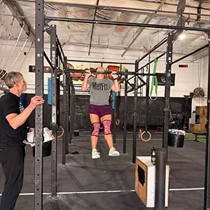 A person with long brown hair in a black long-sleeved shirt and blue jeans stands in a gym near stacks of weight plates.