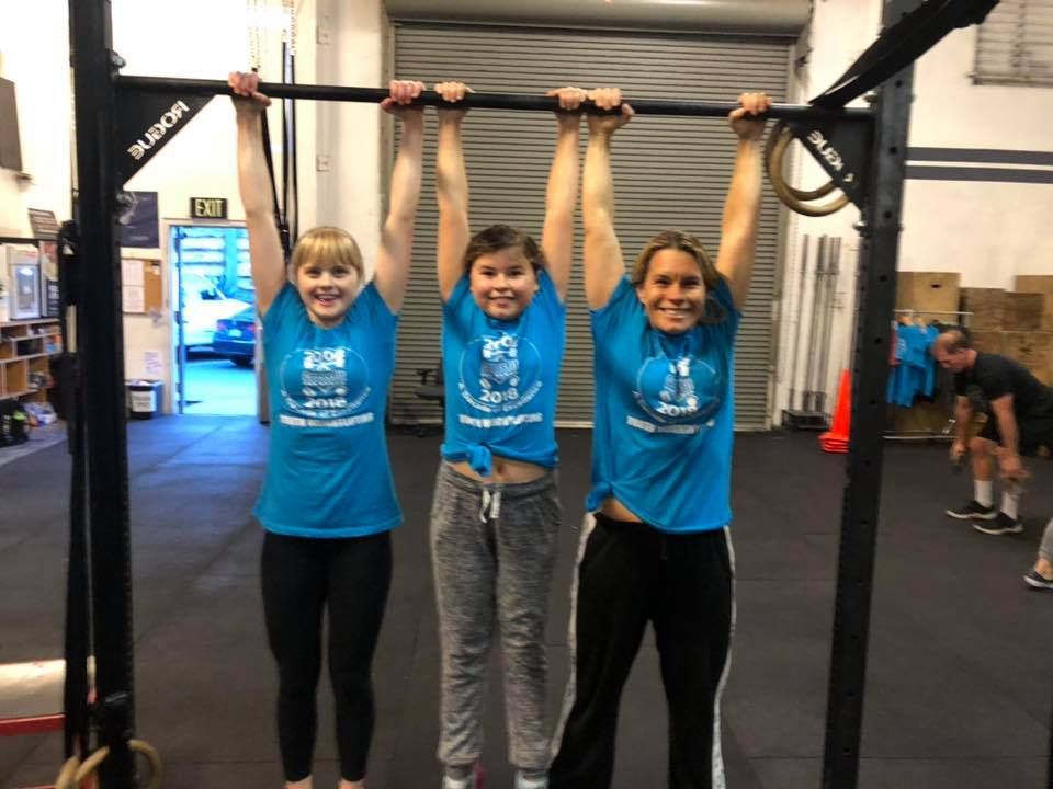 Three people in matching blue shirts hanging from a horizontal metal gym bar inside an indoor fitness facility.