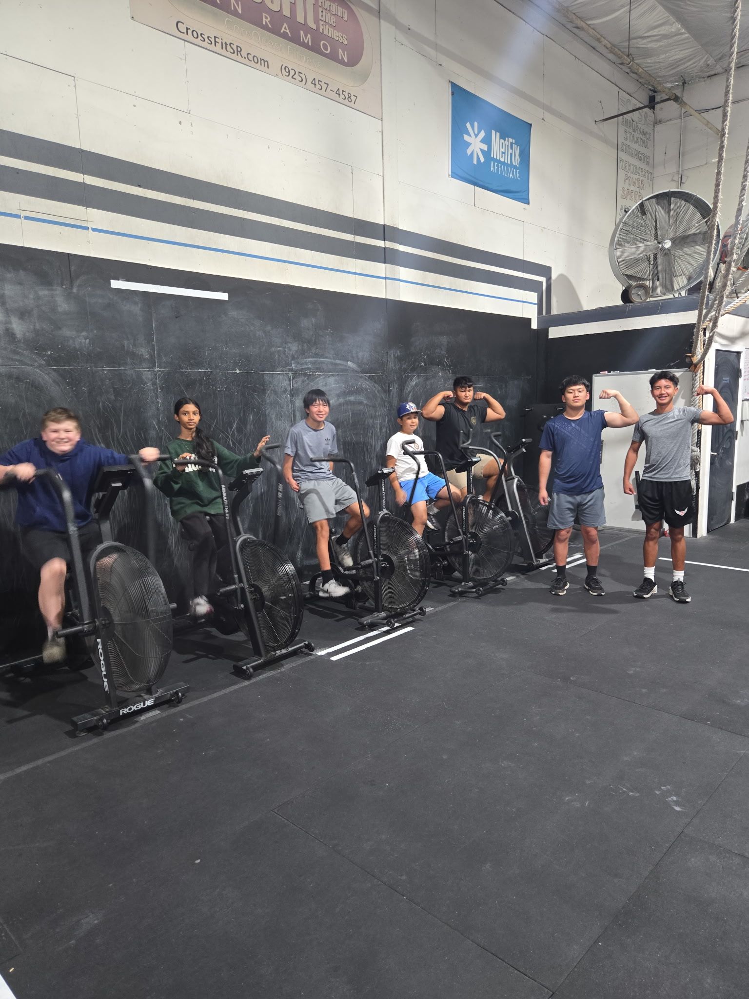 A large, diverse group poses for a photo on an indoor gym's artificial turf, with a barbell on a wooden platform in front.