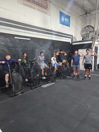 A large, diverse group poses for a photo on an indoor gym's artificial turf, with a barbell on a wooden platform in front.