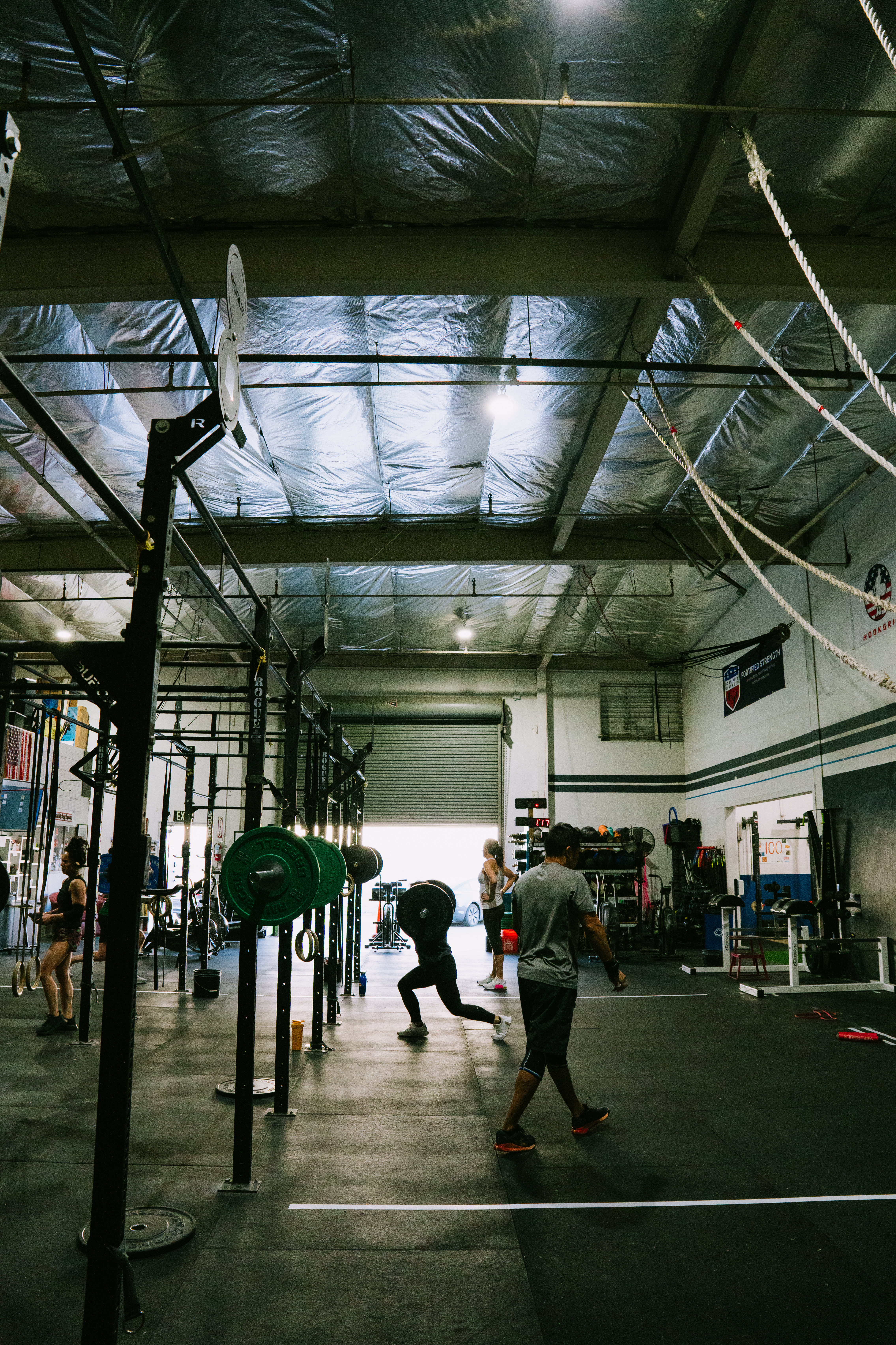 A shirtless person kneeling on a gym mat performs a one-arm row exercise with a red kettlebell in a gym setting.