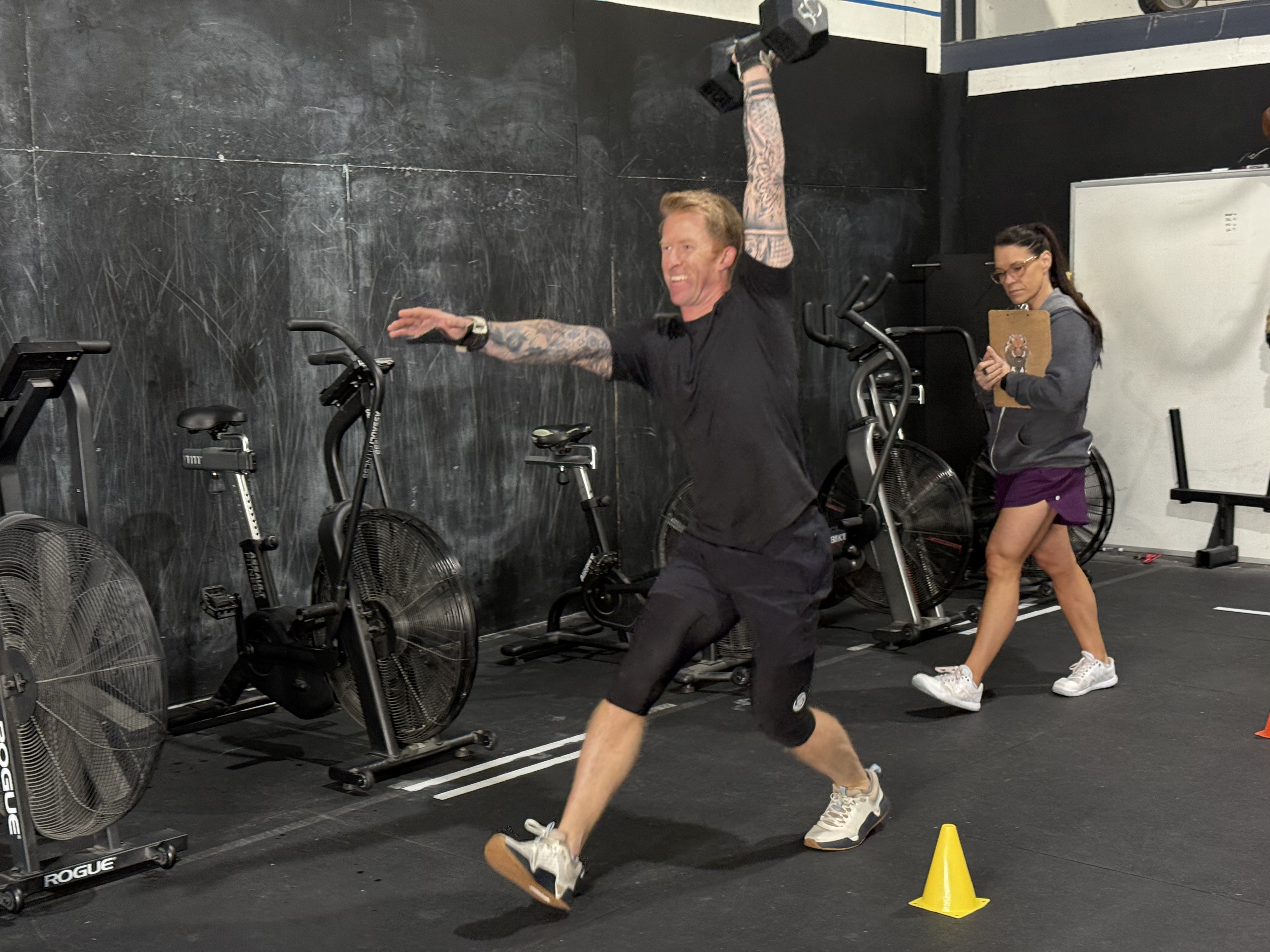 A person with long brown hair in a long-sleeved black shirt talks in a gym setting with stacked weight plates in the back.