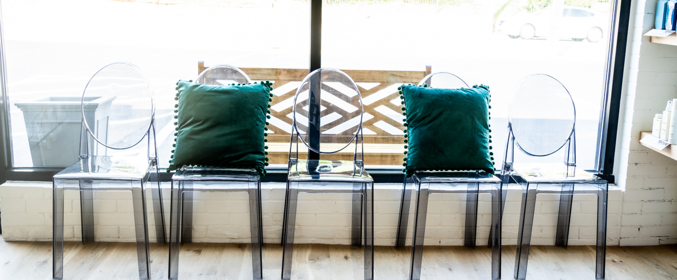 A row of clear chairs with green pillows in front of a window.
