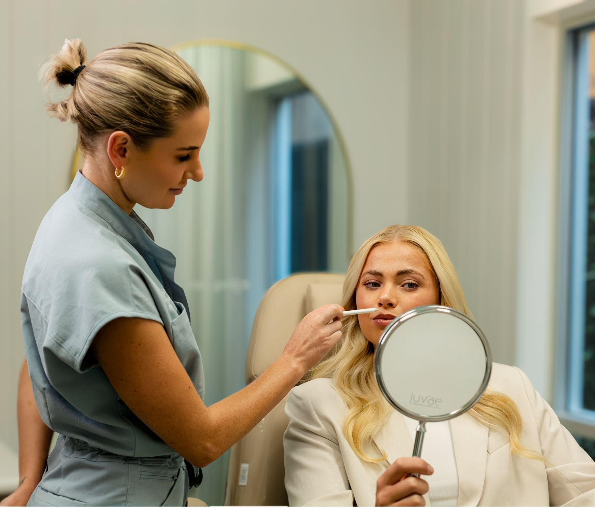 A blonde woman gets lip fillers. A clinician holds a mirror and applies a product.
