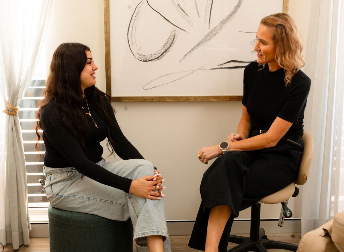 Two women in conversation; one on stool, one in chair. Bright room, art on wall.
