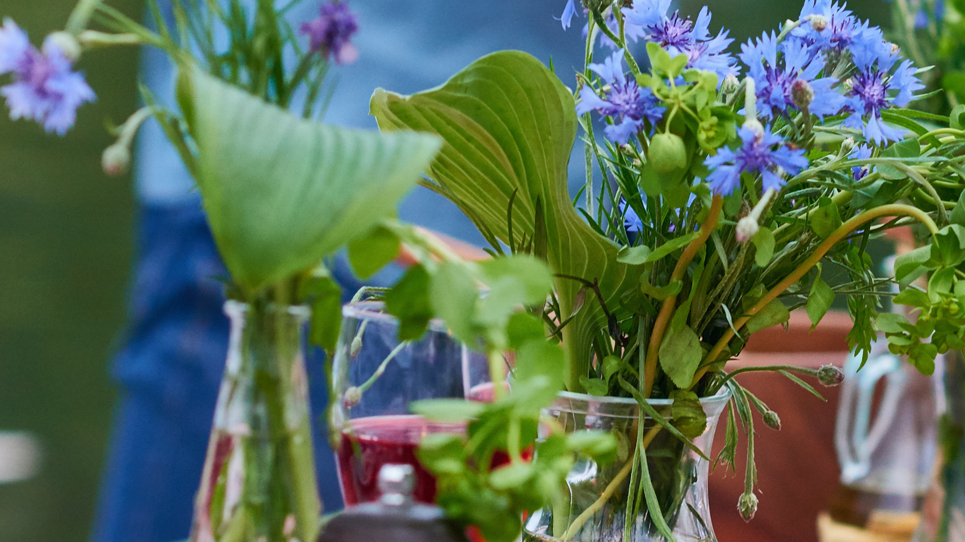 Blaue Blumen in Glasvasen, teilweise mit Grün, neben einem Glas Rotwein auf einem Tisch.