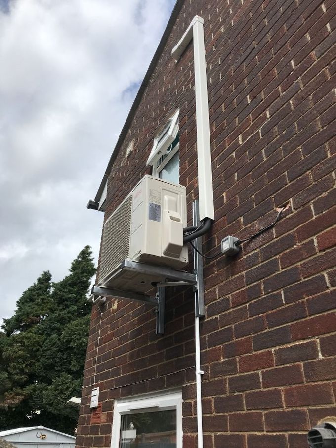 Air conditioning unit mounted on a brick wall with white conduit running up the side.