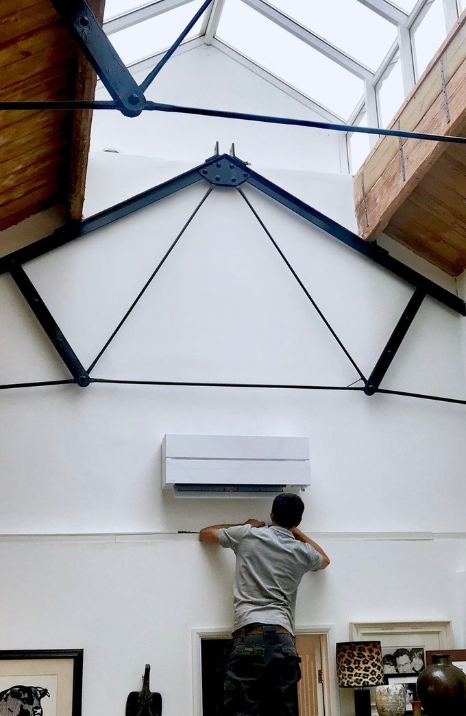 Man on a ladder installing an air conditioning unit on a white wall. Industrial building interior.