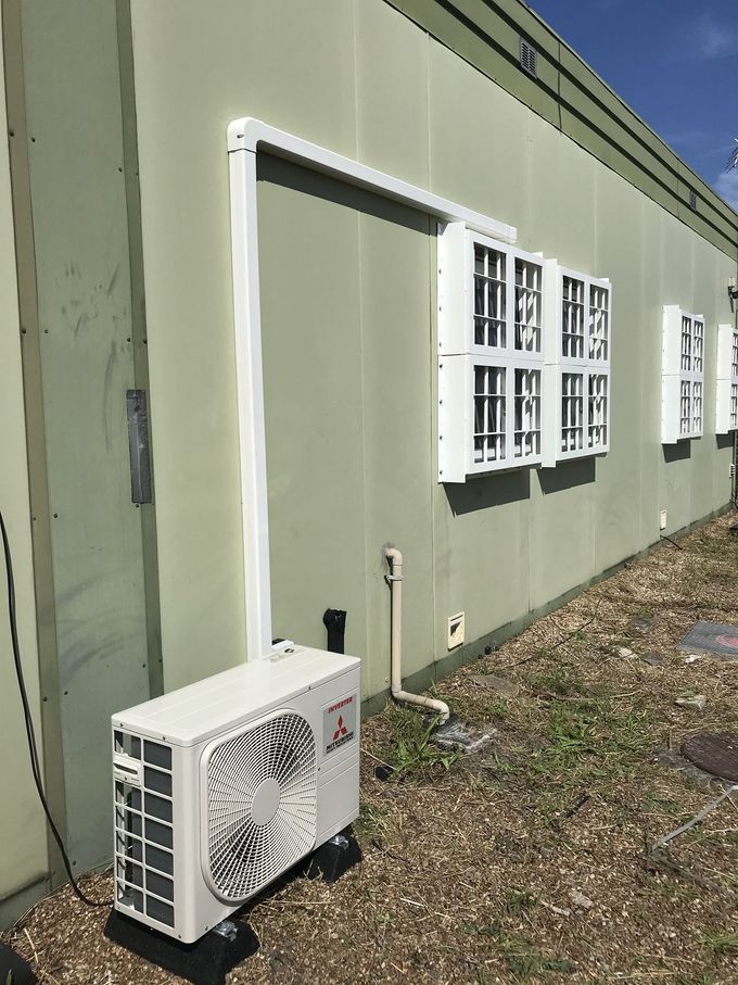 Outdoor HVAC unit next to a light green building. White pipe runs up the wall to the windows.