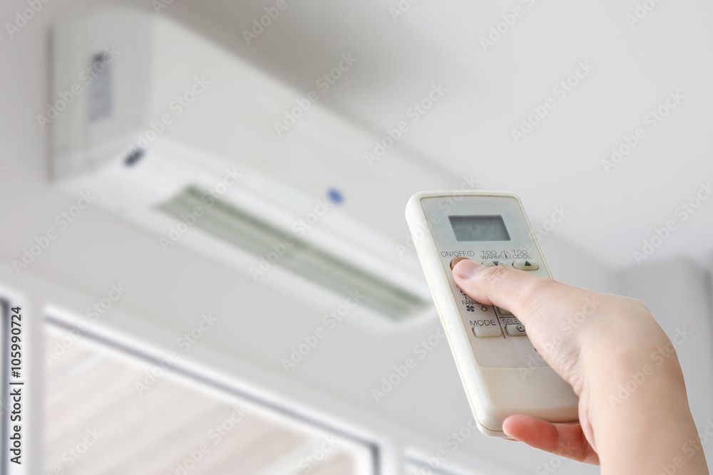 Hand using a remote control to adjust a white air conditioning unit on a white wall.