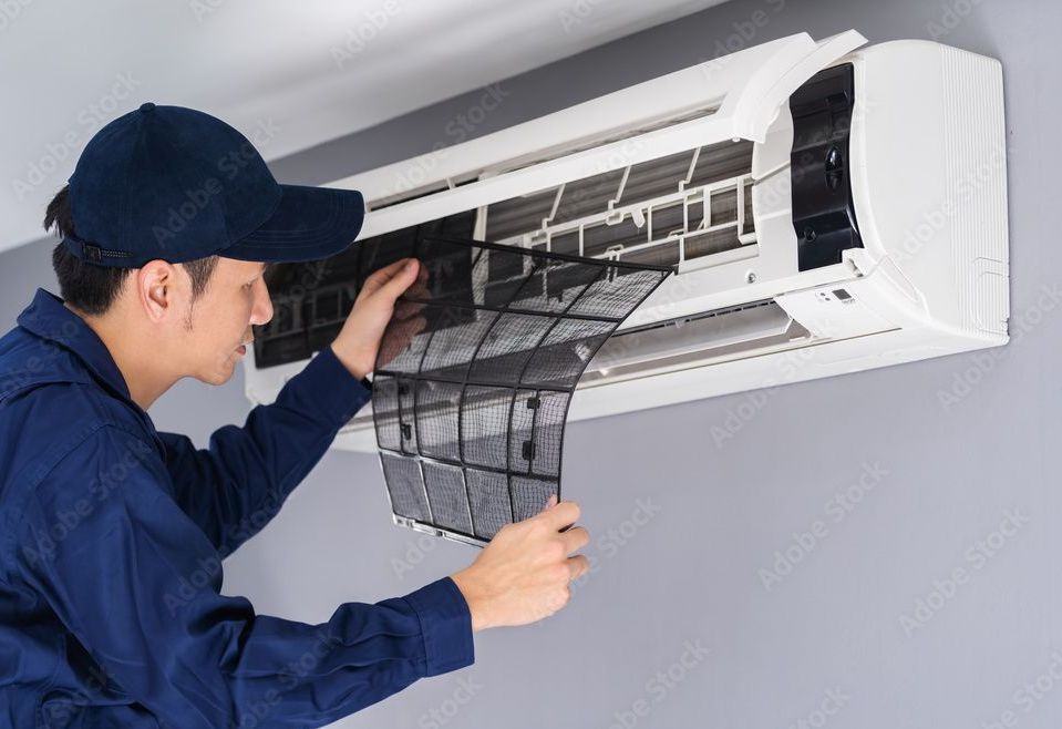 Person in blue uniform removing air filter from an AC unit on a gray wall.