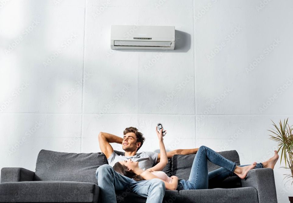 Couple relaxing on couch under air conditioner, man holding remote.