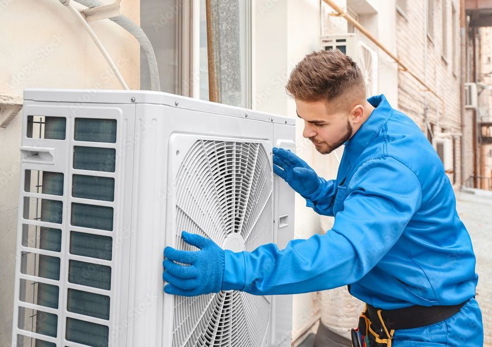 HVAC technician in blue work clothes inspecting an outdoor air conditioning unit.