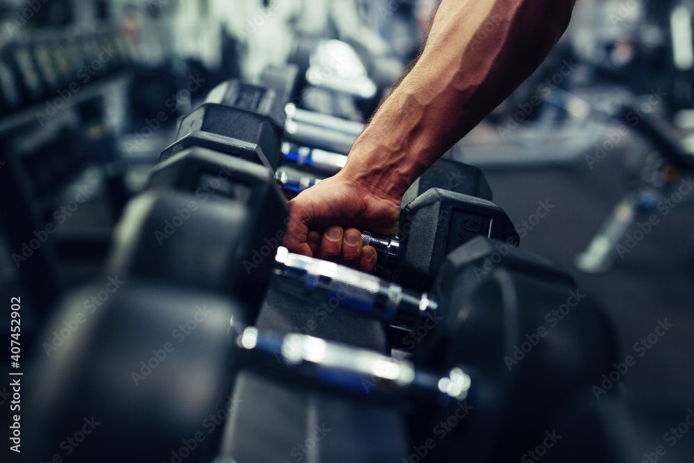 Hand gripping a dumbbell in a gym, arm muscles visible, weights in a rack in the background.
