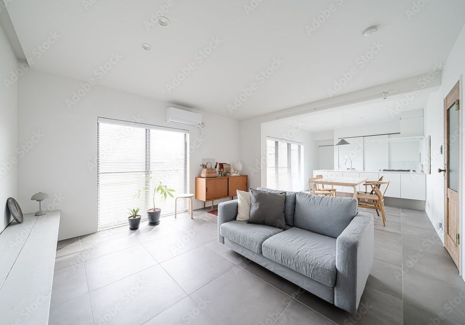 Bright living room with gray sofa, white walls, and a view into a light-filled kitchen.