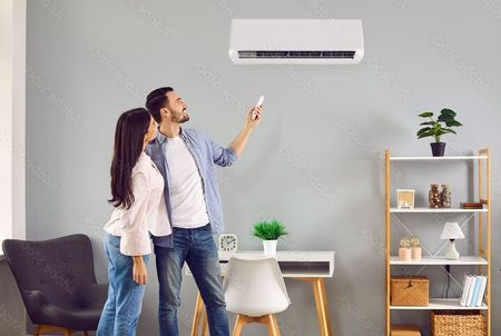 Couple looking up at an air conditioner, man holding remote. Room has desk, shelves, and a chair.