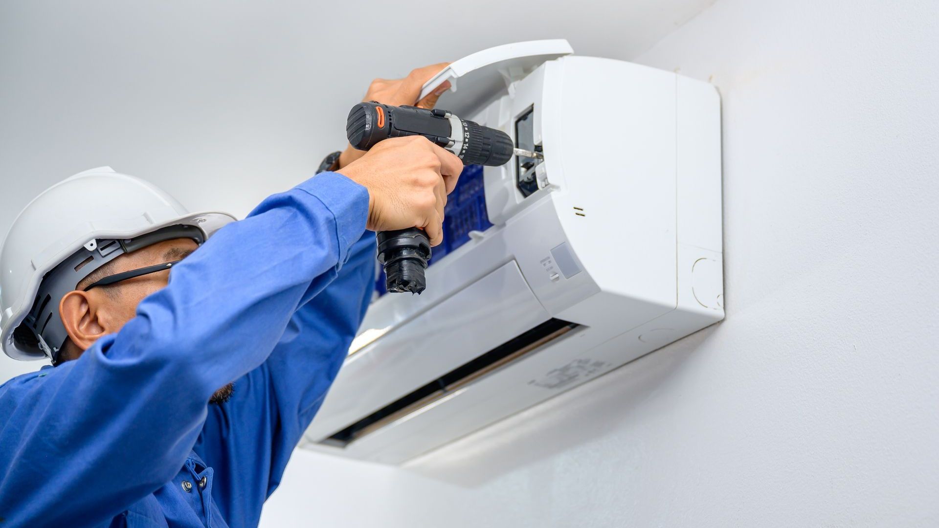 Person in blue jumpsuit installing a wall-mounted air conditioning unit with a power drill.
