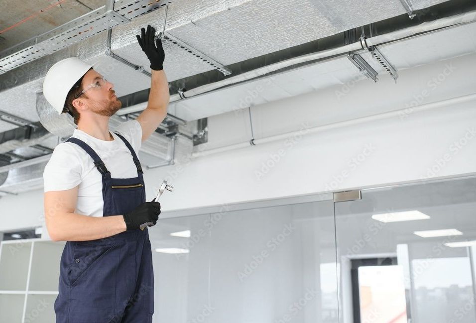Construction worker in white hard hat and blue overalls inspecting ceiling wires.