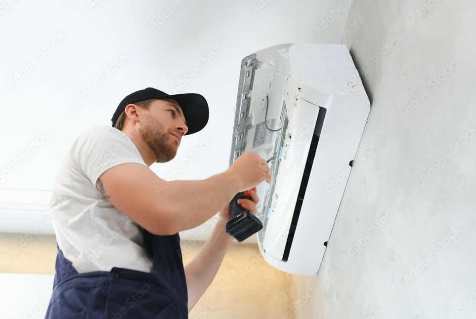 Man in overalls repairs a wall-mounted air conditioner with a power drill indoors.