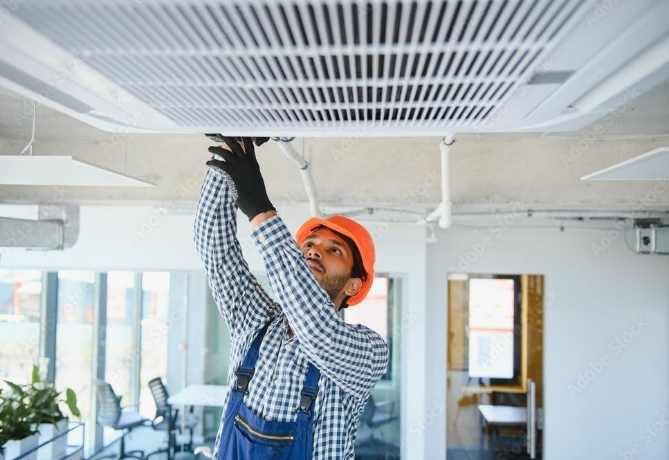 HVAC technician in overalls and hard hat working on a ceiling-mounted air vent in an office.