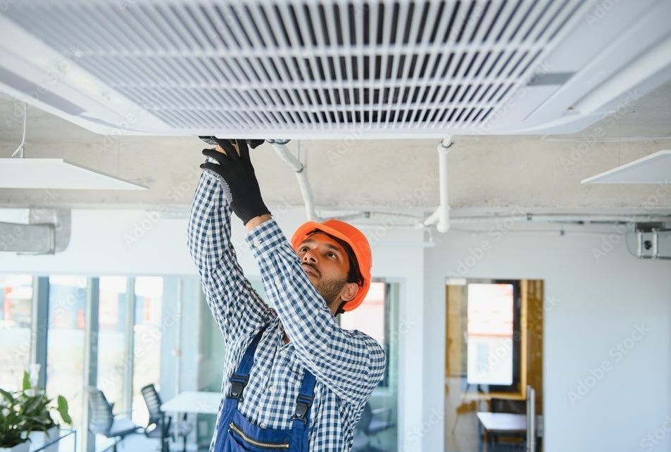 HVAC technician in orange hardhat and gloves, working on ceiling air conditioning unit in an office.