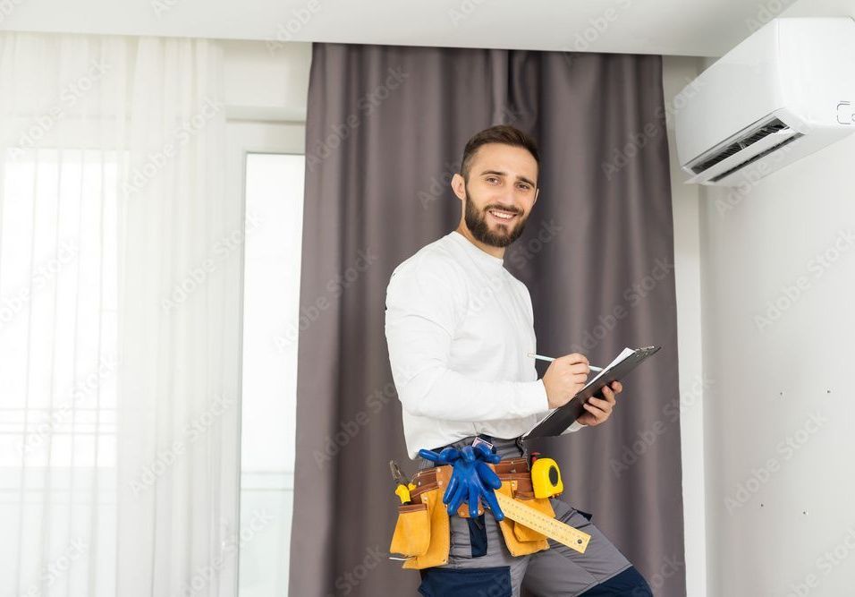 HVAC technician with tool belt, clipboard, inspecting an AC unit indoors.