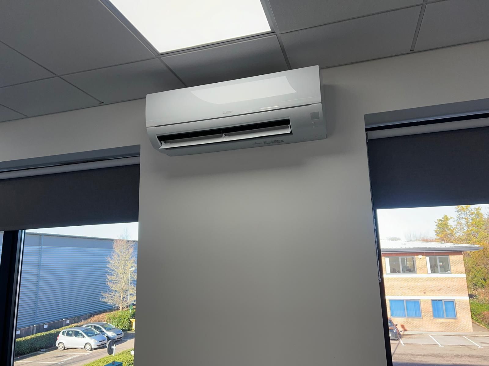 White air conditioning unit mounted on a gray office wall, next to a window with trees and buildings outside.