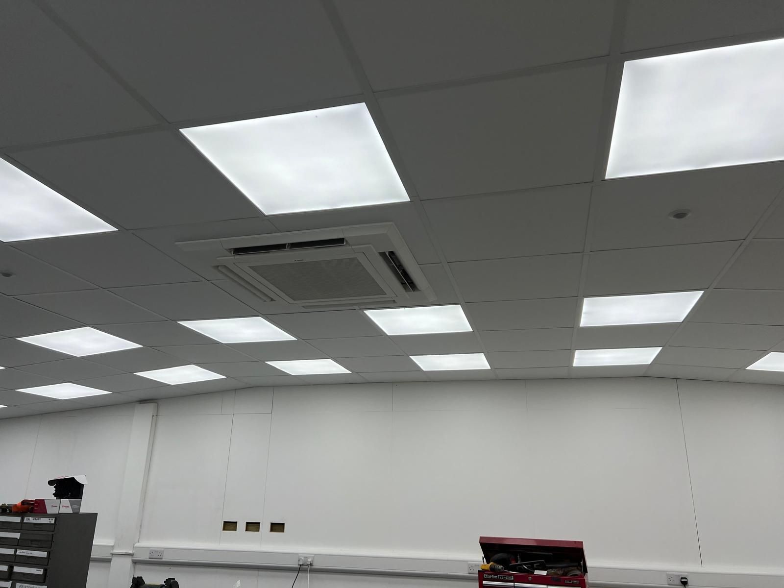 Bright office ceiling with recessed square lights and a central HVAC unit. White walls and workbench visible.
