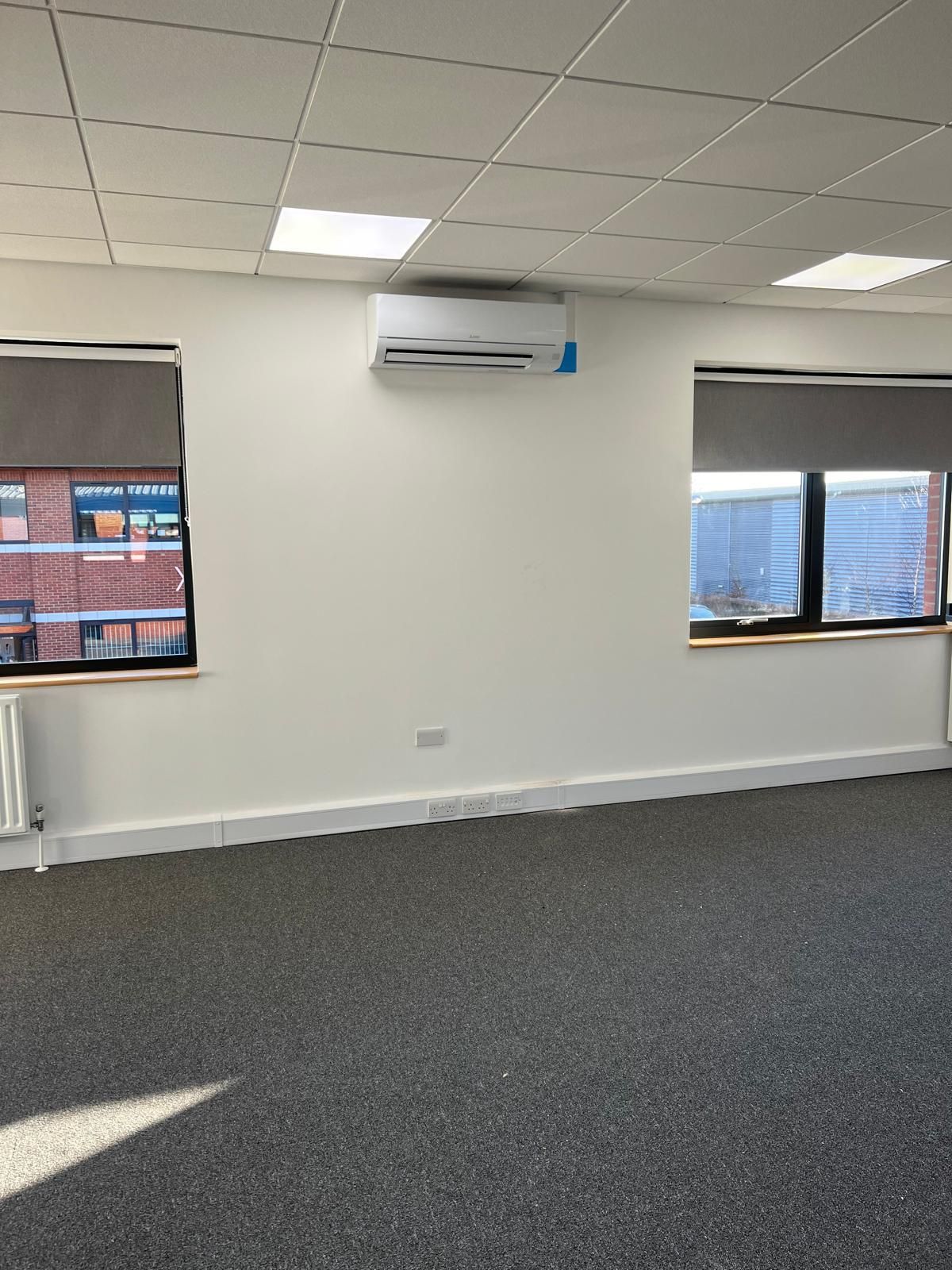 Empty office with gray carpet, white walls, and an air conditioner. Two windows with shades.