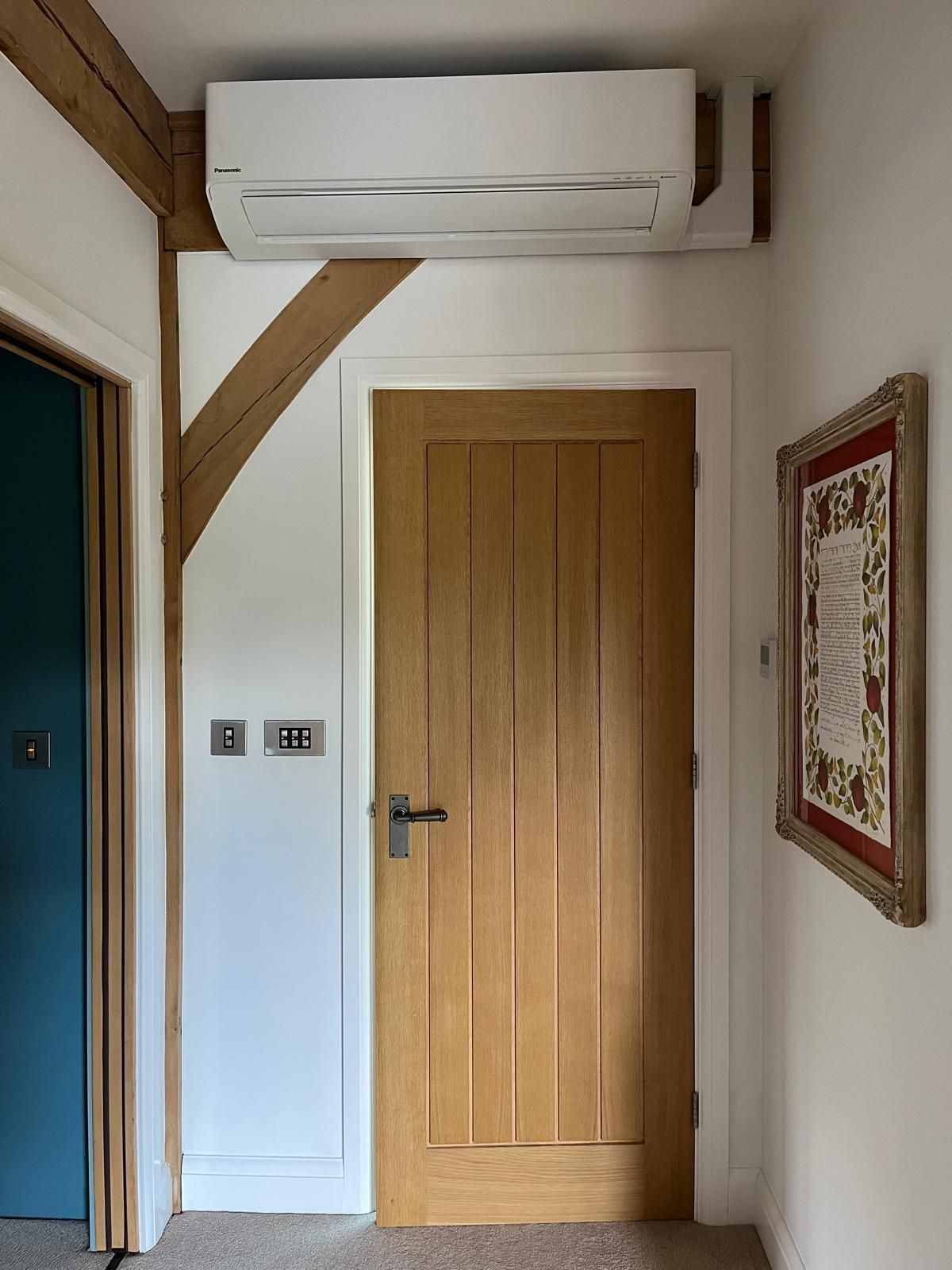 Air conditioning unit above wooden door in a hallway with wood-framed wall and framed artwork.