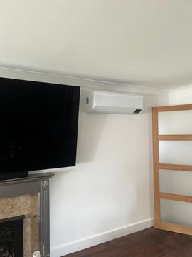 Air conditioner mounted on white wall above a TV and fireplace. Wooden door frame on right.