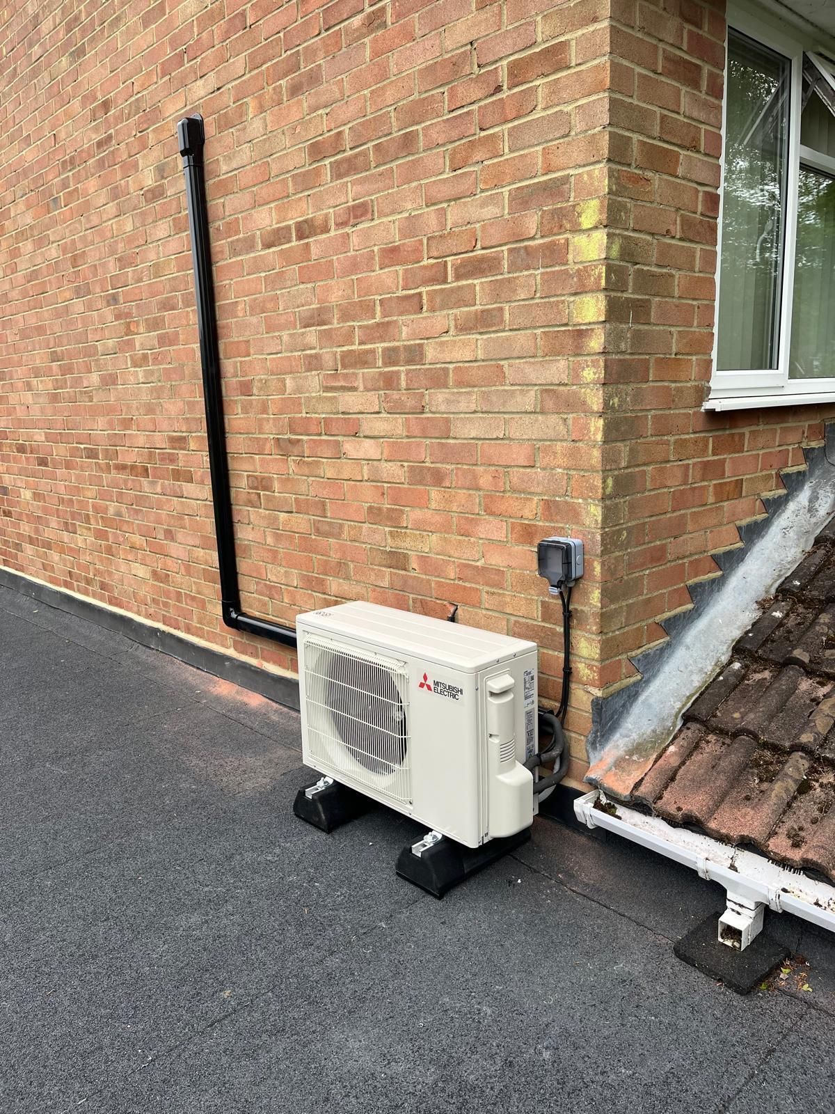 Air conditioning unit on a flat roof, against a brick wall. Black pipe, electrical box, and a window are also visible.