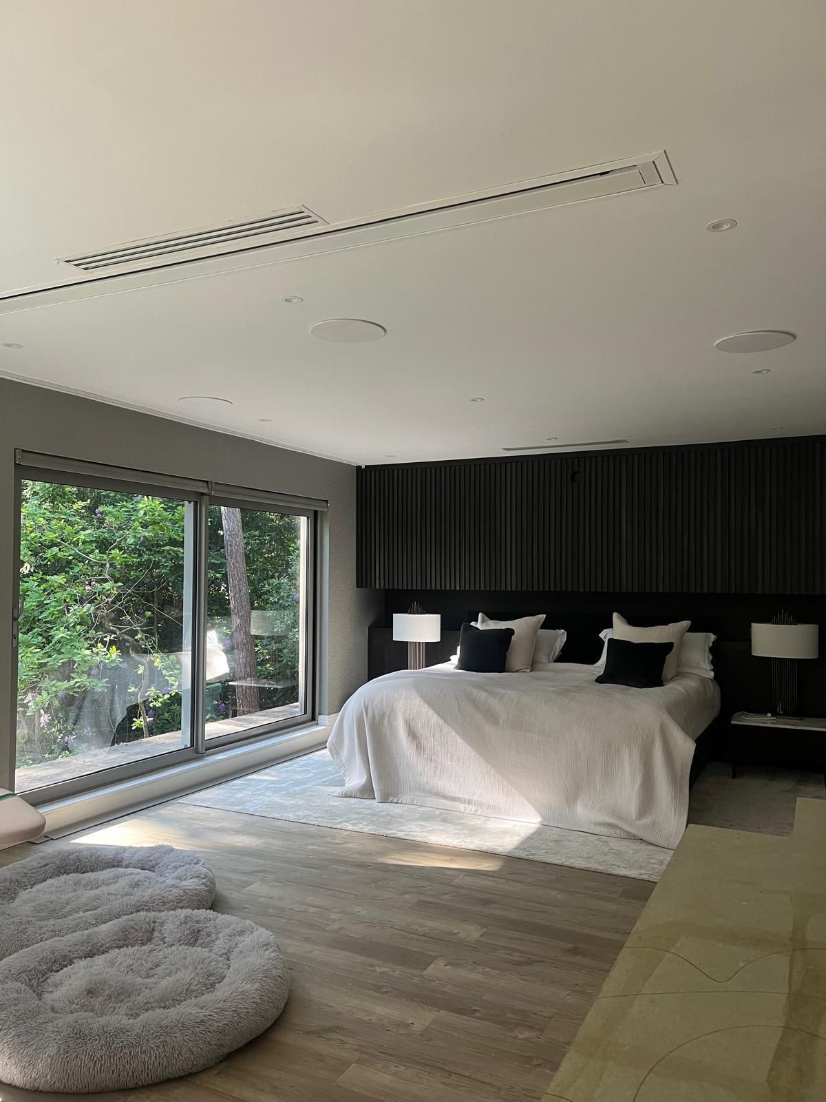 Modern bedroom with large windows, dark headboard, white bedding, and dog beds.
